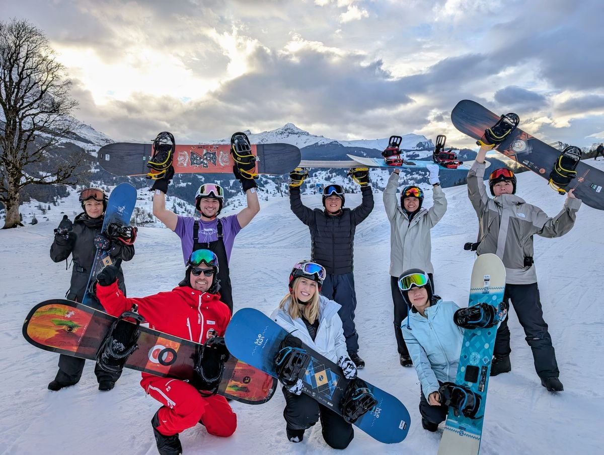 Acht Personen mit Snowboards und Helmen posieren auf schneebedecktem Gelände vor Bergen bei bewölktem Himmel.