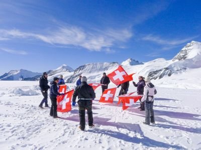 Eine Gruppe mit Schweizer Fahnen auf schneebedecktem Berg, teils mit Winterkleidung und Ausrüstung.