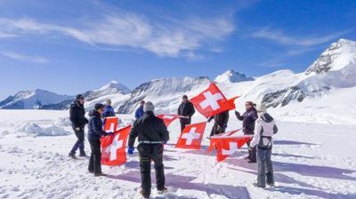 Eine Gruppe mit Schweizer Fahnen auf schneebedecktem Berg, teils mit Winterkleidung und Ausrüstung.