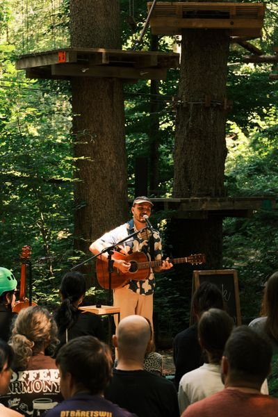 Person mit Helm und Gitarre spielt vor Publikum im Wald, Kletterpark mit Seilbrücken im Hintergrund.