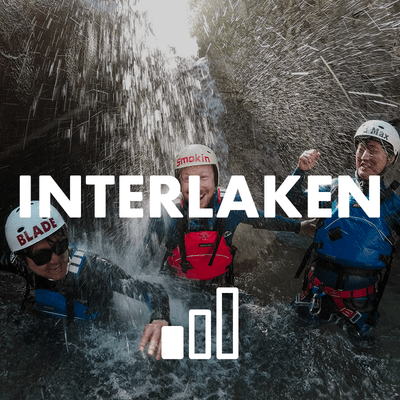 Drei Personen in Helmen und Neoprenanzügen stehen unter einem Wasserfall in einer felsigen Canyon-Umgebung.