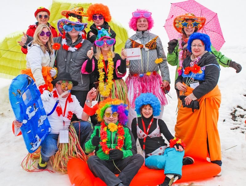 A group in colorful costumes and wigs is posing on snow with sleds and umbrellas.
