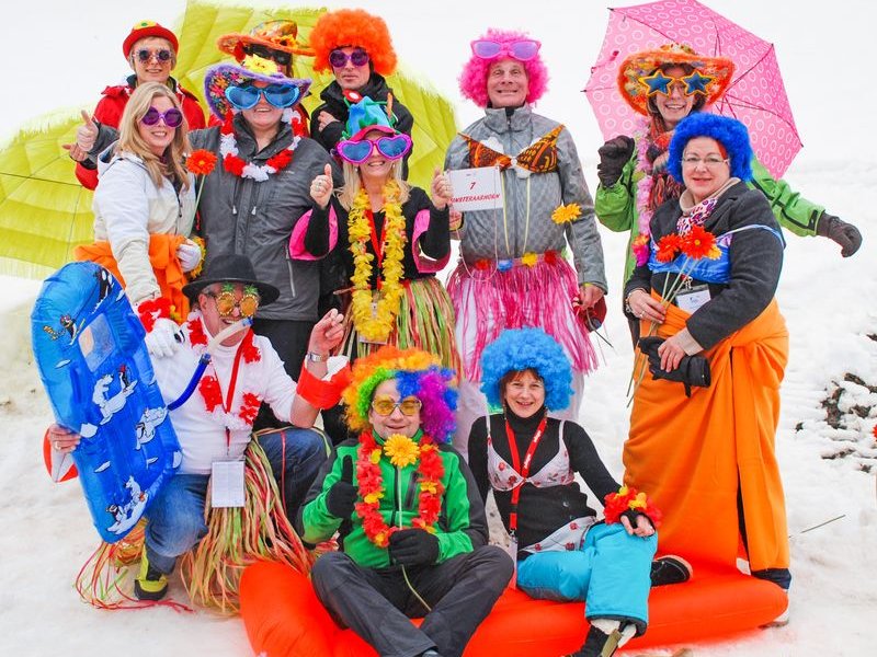 A group in colorful costumes and wigs is posing on snow with sleds and umbrellas.