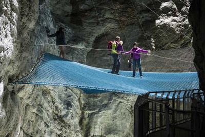 People on a blue net in a rocky gorge, secured with ropes, wearing casual clothing.