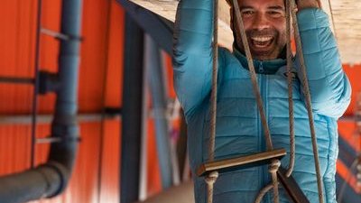 Person in blue jacket and helmet climbing a rope ladder indoors, using safety gear and harness.