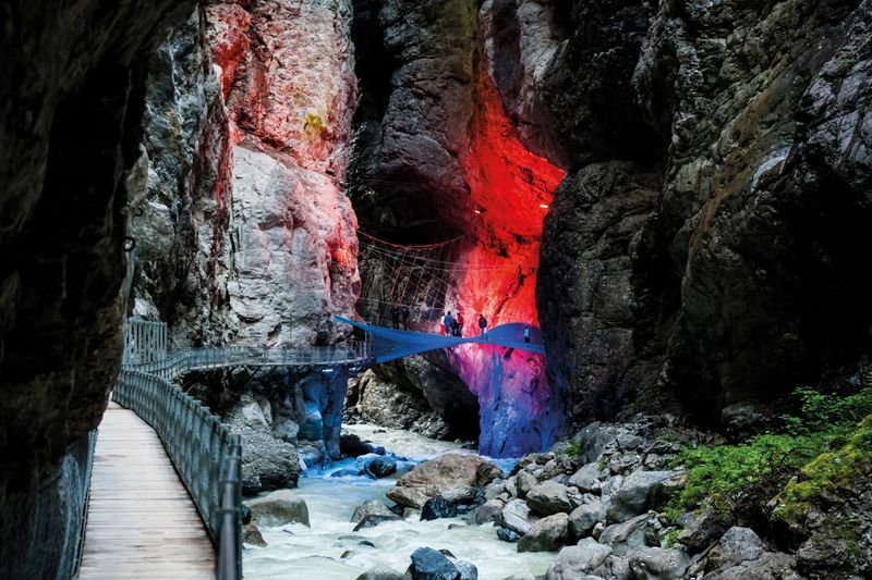 People on an illuminated walkway in a gorge, surrounded by rocks and flowing water.