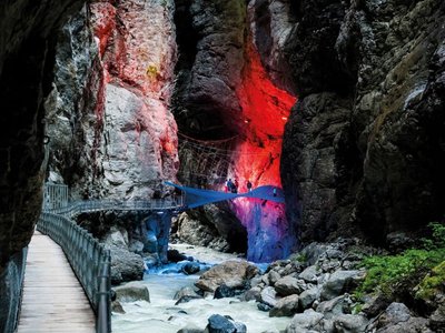 People on an illuminated walkway in a gorge, surrounded by rocks and flowing water.
