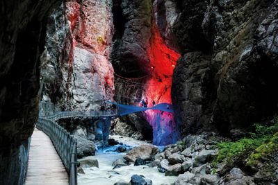 Personen auf einem beleuchteten Steg in einer Schlucht, umgeben von Felsen und fließendem Wasser.