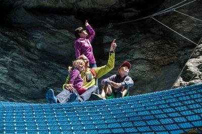 People on blue net in a rocky landscape, wearing casual clothing, pointing upwards.