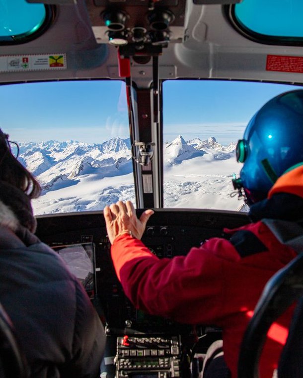 Two people in a helicopter cockpit, one wearing a blue helmet, flying over snowy mountains.