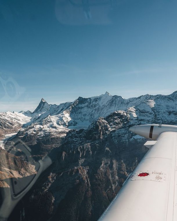 View from airplane wing over snow-capped mountains under clear blue sky.