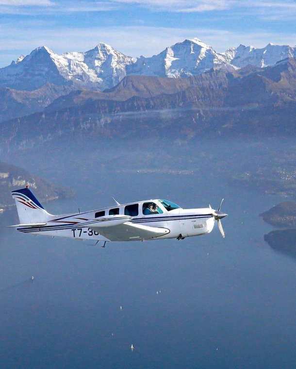 Small airplane flying over a lake with snow-capped mountains in the background.