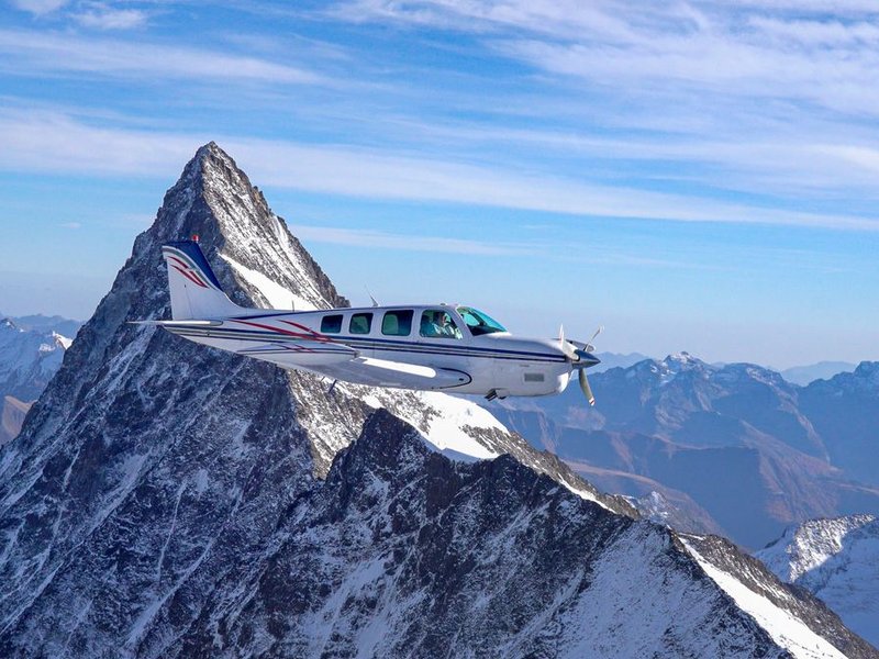 Small airplane flying past a snow-covered mountain peak under a clear blue sky.