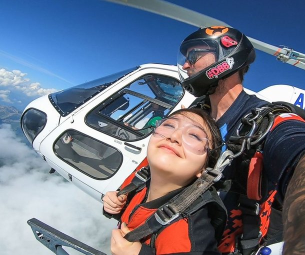 Zwei Personen, die aus einem Hubschrauber Fallschirmspringen, tragen Helme und Gurte, über Wolken und Bergen.
