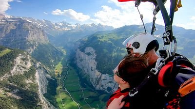 Two people paragliding over a green valley with mountains, wearing helmets and harnesses.