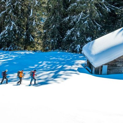 Drei Personen machen Schneeschuhwandern in der Nähe einer schneebedeckten Hütte in einem bewaldeten Gebirgsgebiet und tragen Rucksäcke.