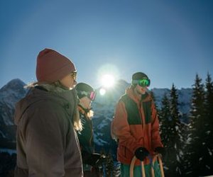 Drei Personen in Winterkleidung mit Schlitten, die Skibrillen und Mützen tragen, in einer verschneiten Berglandschaft.