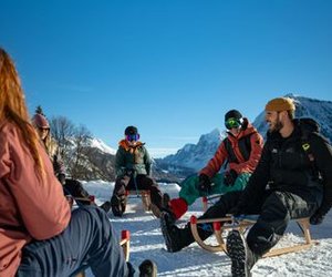 People sitting on sleds in snowy mountains, wearing winter clothing and goggles, clear blue sky.
