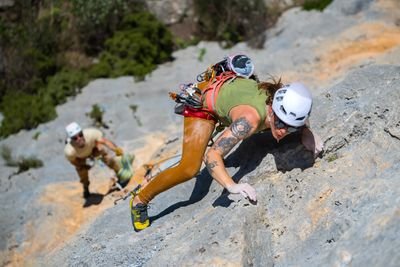 Two climbers with helmets and harnesses ascend a rocky cliff, surrounded by greenery.