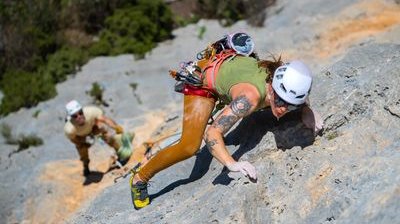 Two climbers with helmets and harnesses ascend a rocky cliff, surrounded by greenery.