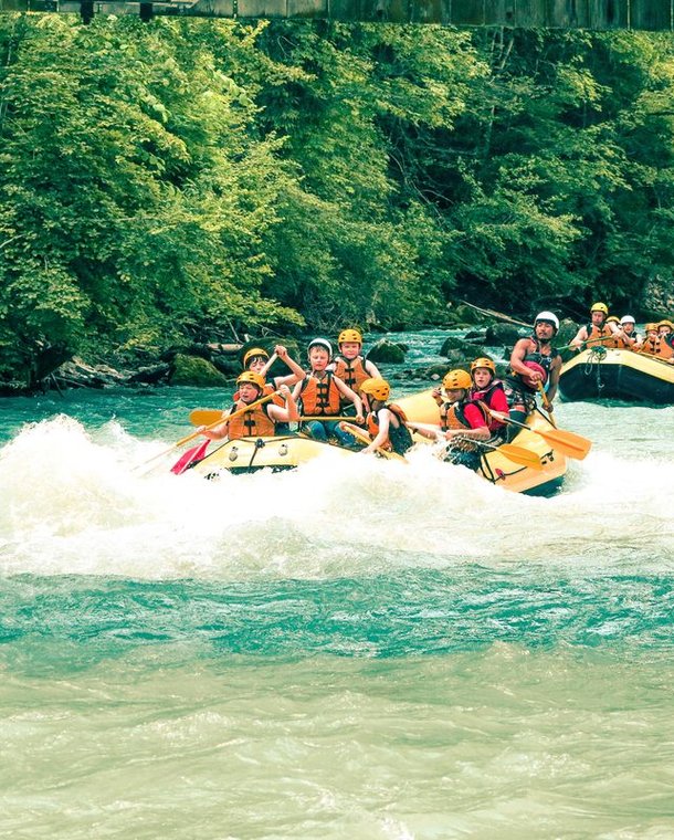 Groups rafting on a river, wearing helmets and life jackets, surrounded by lush green trees.