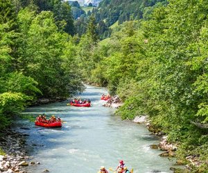 People in helmets and life jackets rafting on the Simme river, surrounded by lush green forests and mountains.