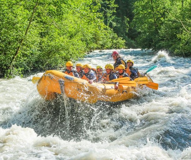 Group in yellow helmets and life jackets rafting on the Simme River, surrounded by trees.