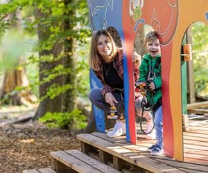 Kind mit Sicherheitsgurt, begleitet von erwachsener Person auf einer hölzernen Plattform im Seilpark Interlaken im Wald.