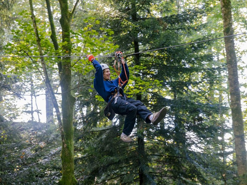 Person riding a zipline through the forest is wearing a helmet, a safety harness, and gloves, surrounded by trees.