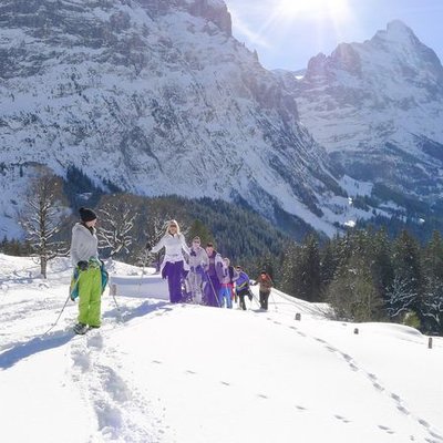 Personen in bunter Winterkleidung wandern im Schnee vor einer Bergkulisse.