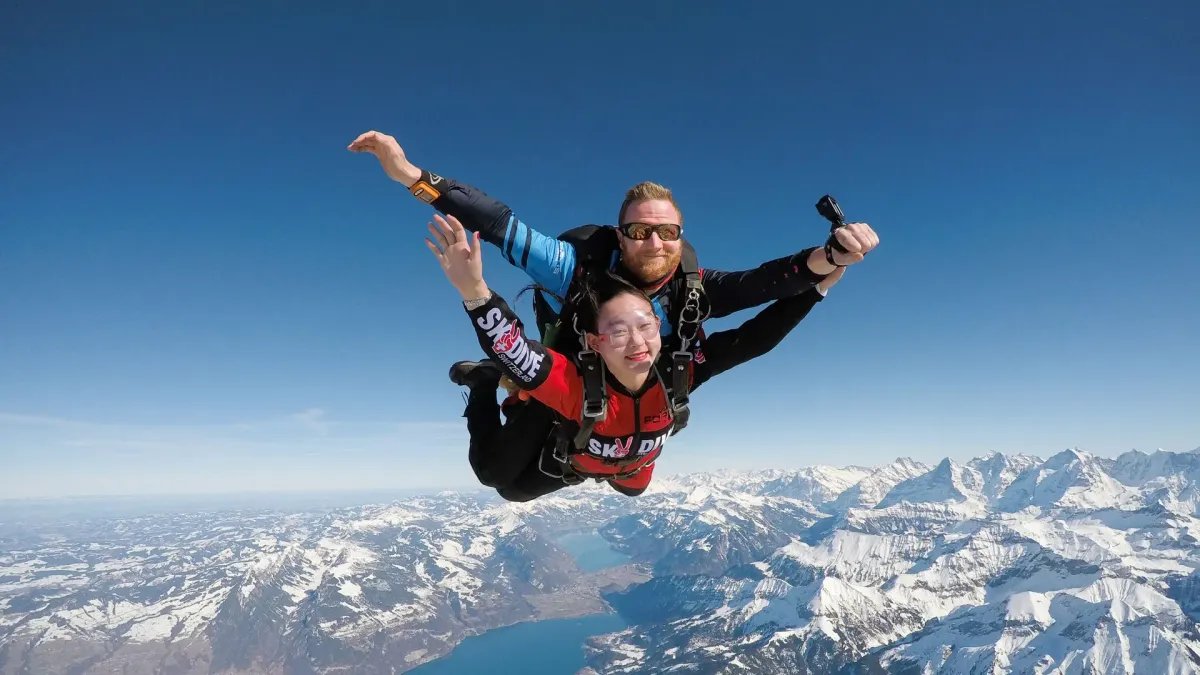 Zwei Personen beim Tandem-Fallschirmsprung mit Helmen und Anzügen über verschneiten Alpen.