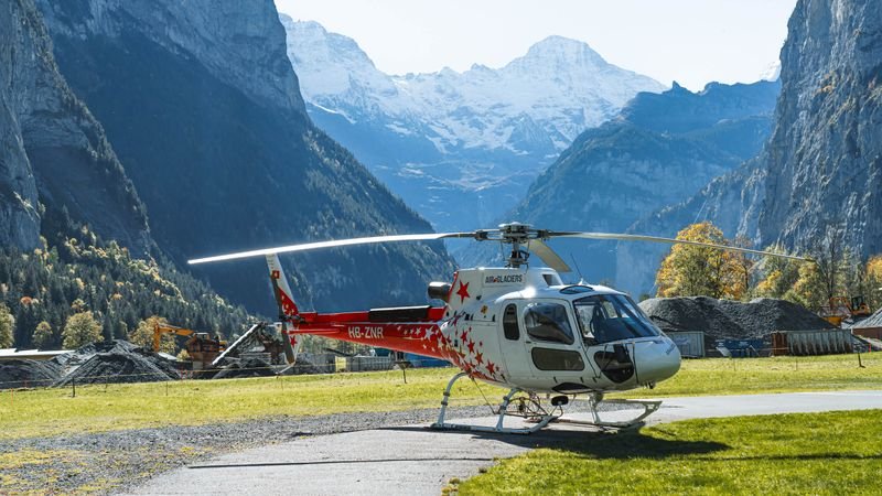 Ein roter Helikopter steht auf einer Wiese in einem alpinen Tal mit hohen Bergen im Hintergrund.