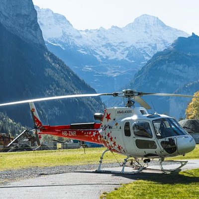 Ein roter Helikopter steht auf einer Wiese in einem alpinen Tal mit hohen Bergen im Hintergrund.