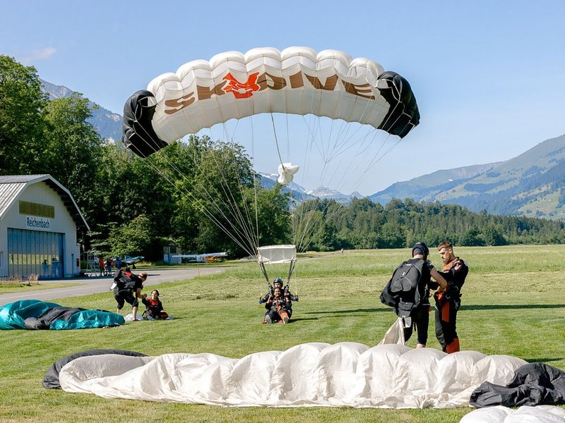 Parachutists land on a meadow in front of a hangar in the Swiss Alps, with parachutes.