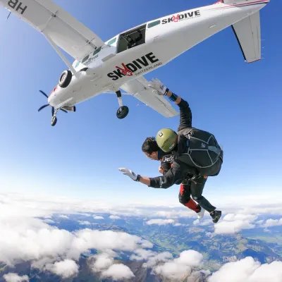 Fallschirmspringer mit Helm und Gurtzeug im Tandemsprung aus Flugzeug über Berglandschaft.
