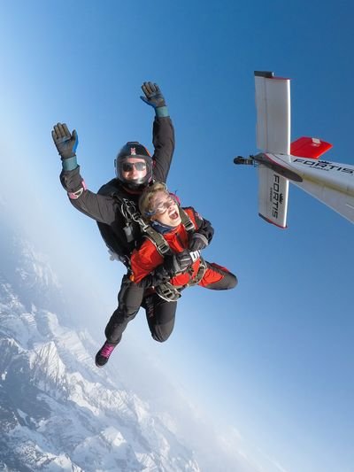 Tandem-Fallschirmspringer im freien Fall über schneebedeckten Bergen, tragen Helme und Gurte, in der Nähe eines Flugzeugs.