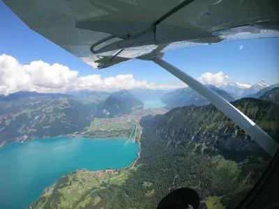 Blick aus einem Flugzeug auf einen türkisfarbenen See, umgeben von Bergen und Wäldern.