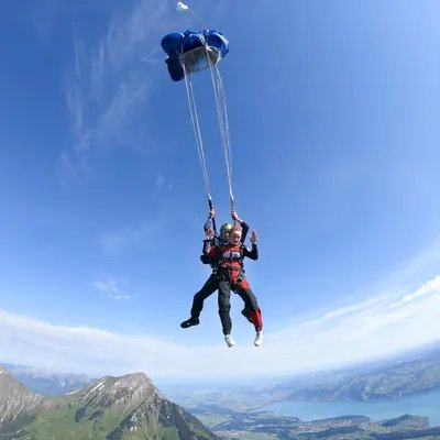 Zwei Personen beim Tandem-Fallschirmsprung über Bergen, mit blauen Fallschirmen und Sicherheitsausrüstung.