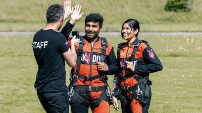 Two people in skydiving suits with harnesses on a meadow, accompanied by a staff member.